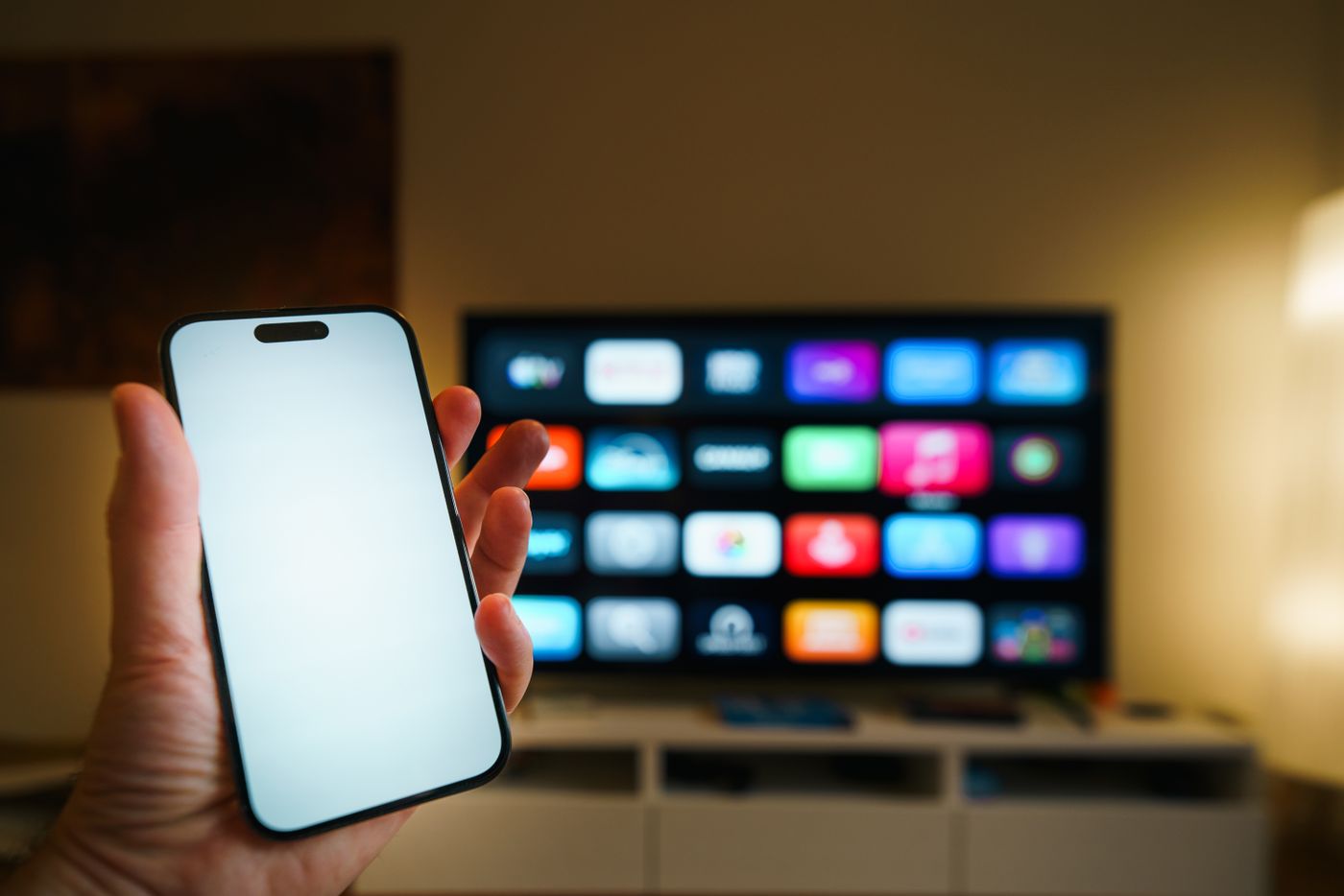 A smartphone in the foreground with a smart TV showing its app menu in a softly lit living room